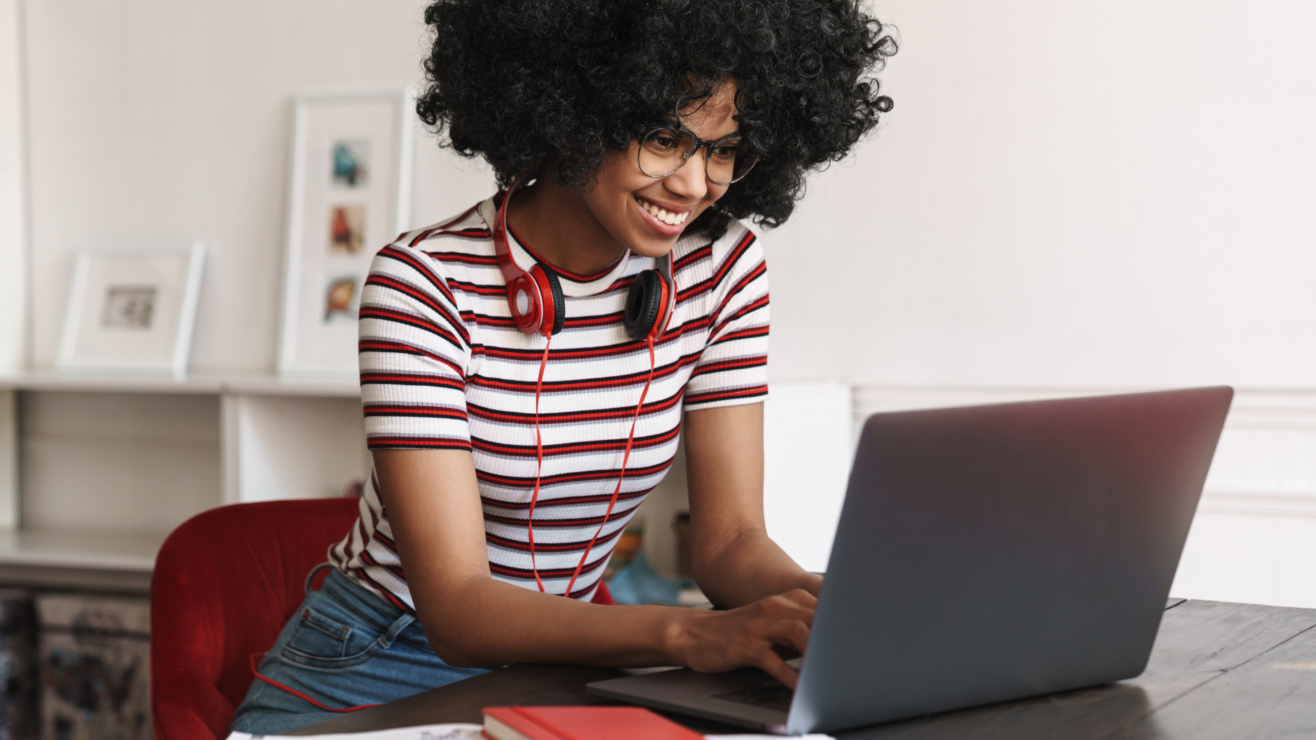 A woman sits at a table, smiling while looking at her laptop and typing.